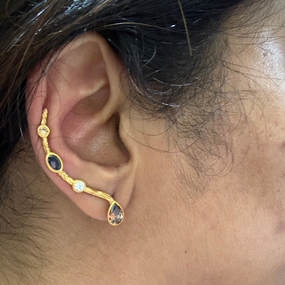 A close-up of a person's ear wearing a gold ear cuff earring with three dark oval stones, a small clear round stone, and a teardrop-shaped clear stone.