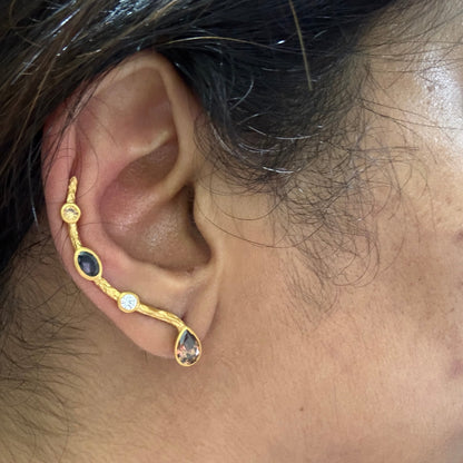 A close-up of a person's ear wearing a gold ear cuff earring with three dark oval stones, a small clear round stone, and a teardrop-shaped clear stone.