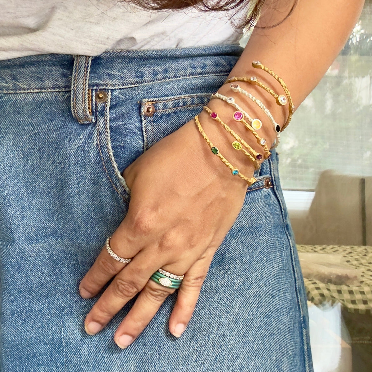 A close-up of a person wearing multiple delicate gold bracelets and rings, with one bracelet featuring colorful gemstone accents. The person is wearing blue denim jeans and a light gray top, with their hand resting in their pocket.