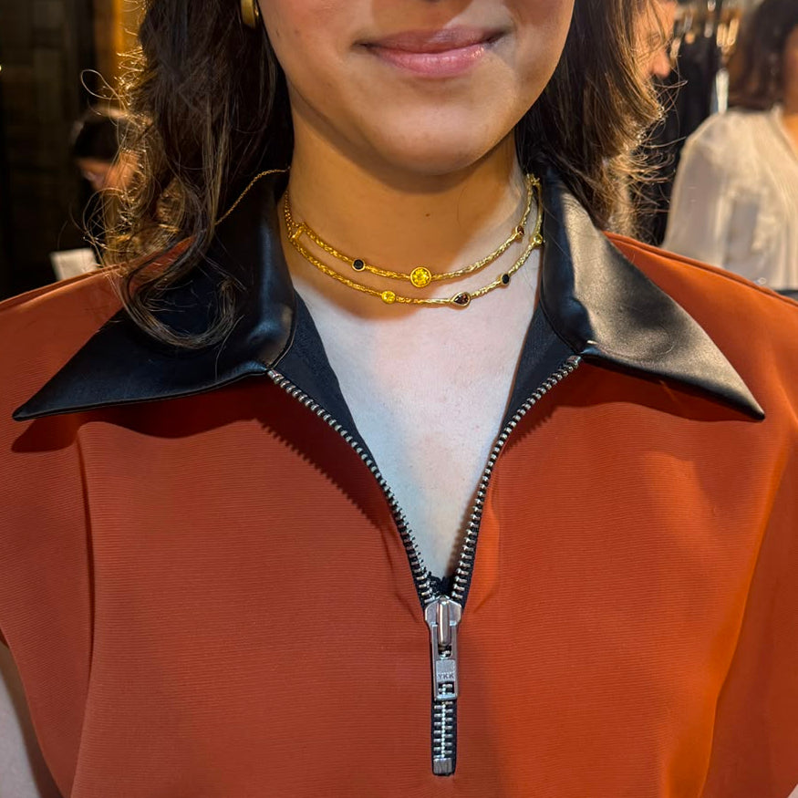 A woman wearing a sleeveless rust-orange top with a black collar and a front zipper, photographed in a backstage or event setting with clothing racks and bright overhead lights in the background.