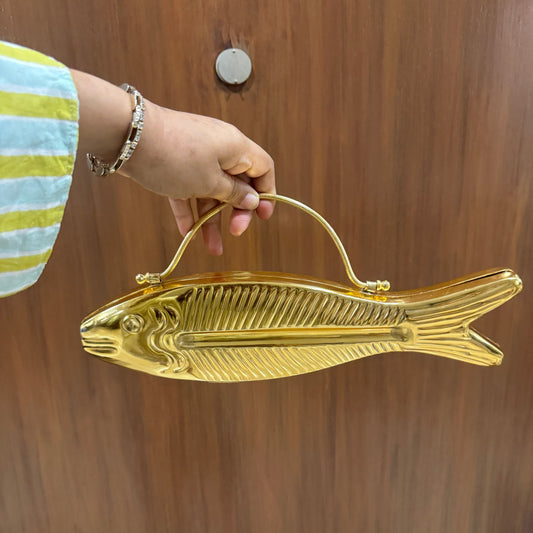 A hand holding a shiny gold fish-shaped handbag with a top handle, set against a brown wooden door background.