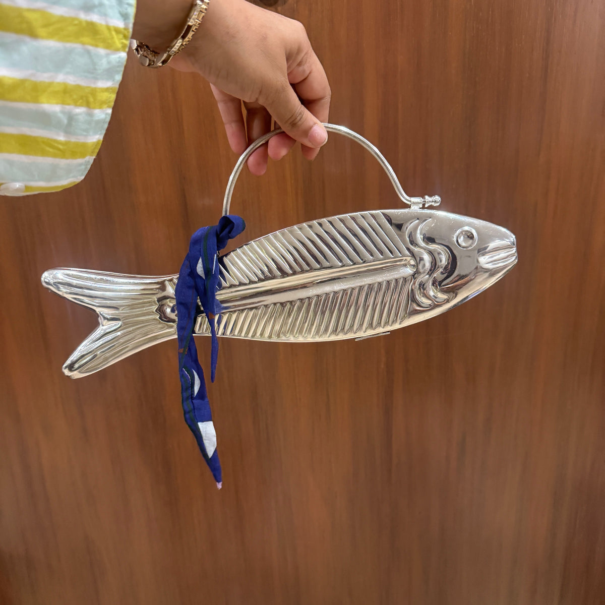 A hand holding a silver fish-shaped handbag with a top handle and a blue ribbon tie, set against a brown wooden door background.