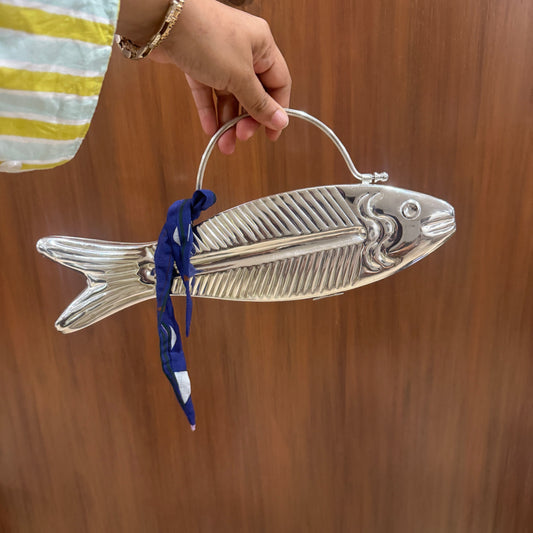 A hand holding a silver fish-shaped handbag with a top handle and a blue ribbon tie, set against a brown wooden door background.
